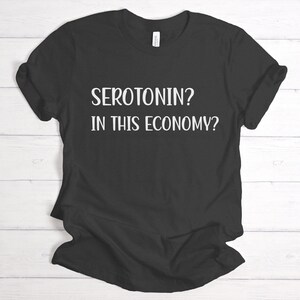 Pu&ograve; includere: Maglietta nera con testo bianco che recita "Serotonina? In questa economia?"
