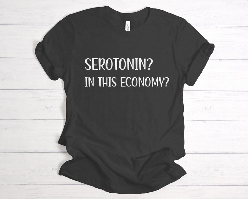 Pu&ograve; includere: Maglietta nera con testo bianco che recita "Serotonina? In questa economia?"