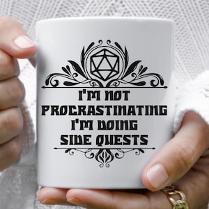 May include: White ceramic mug with a black design featuring a 20-sided die, a floral design, and the text "I'm not procrastinating I'm doing side quests".
