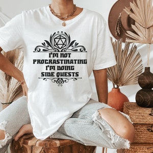 May include: White t-shirt with the text "I'M NOT PROCRASTINATING I'M DOING SIDE QUESTS" in a black decorative font. The design includes a geometric shape and flourishes. The shirt is a casual wear item.