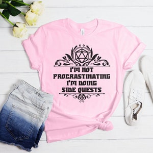 May include: A light pink t-shirt with the text "I'M NOT PROCRASTINATING I'M DOING SIDE QUESTS" in black. The design includes a geometric shape and decorative flourishes. The shirt is paired with denim shorts and white sneakers.