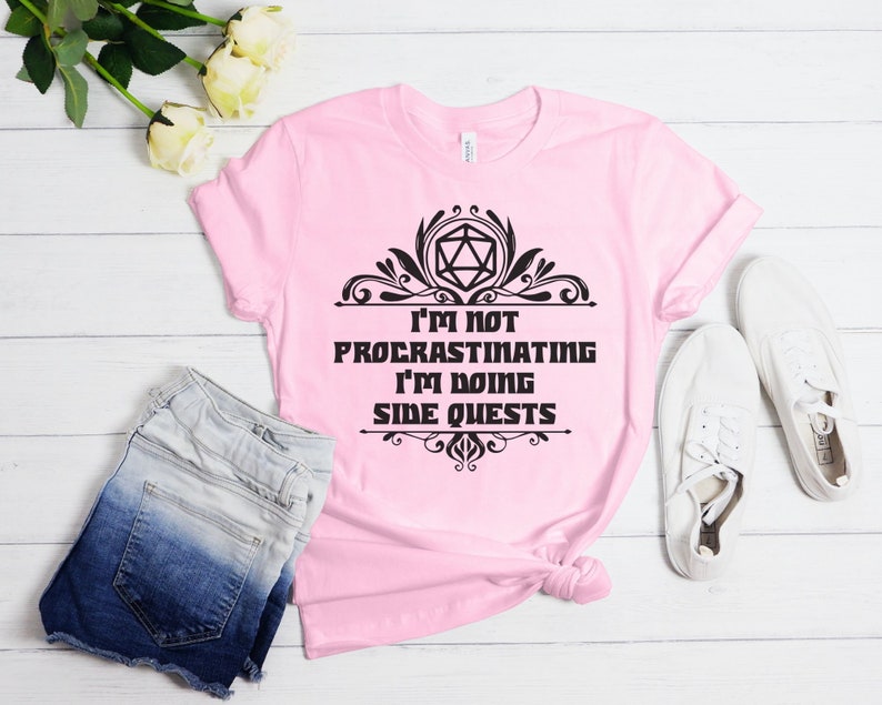 May include: A light pink t-shirt with the text "I'M NOT PROCRASTINATING I'M DOING SIDE QUESTS" in black. The design includes a geometric shape and decorative flourishes. The shirt is paired with denim shorts and white sneakers.