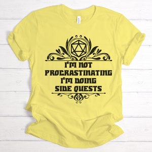 May include: A yellow t-shirt with the text "I'M NOT PROCRASTINATING I'M DOING SIDE QUESTS" in black. The text is framed by decorative black flourishes and a geometric shape. The shirt is made of a soft material.