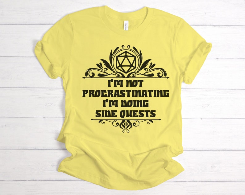 May include: A yellow t-shirt with the text "I'M NOT PROCRASTINATING I'M DOING SIDE QUESTS" in black. The text is framed by decorative black flourishes and a geometric shape. The shirt is made of a soft material.