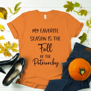 Op de afbeelding: Oranje T-shirt met de tekst "My favorite season is the Fall of the Patriarchy" in zwart gedrukt.