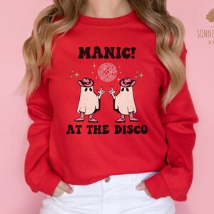 May include: Red sweatshirt with a white graphic of two ghosts wearing cowboy hats and dancing in front of a disco ball. The text "MANIC! AT THE DISCO" is printed in black below the ghosts.