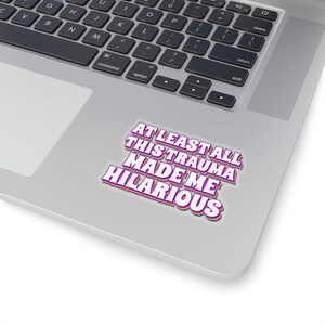 May include: A sticker with the text "At least all this trauma made me hilarious" in a pink, green, and white retro font on a silver laptop.
