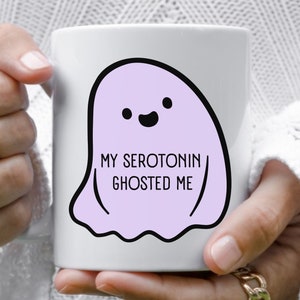 May include: White ceramic mug featuring a purple ghost illustration and the text "My Serotonin Ghosted Me".