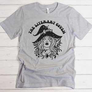 Peut inclure: Un t-shirt gris avec un motif noir d'une sorci&egrave;re portant un chapeau orn&eacute; d'&eacute;toiles et d'un croissant de lune. Le texte "The Literary Coven" est &eacute;crit dans une police r&eacute;tro autour de la sorci&egrave;re.