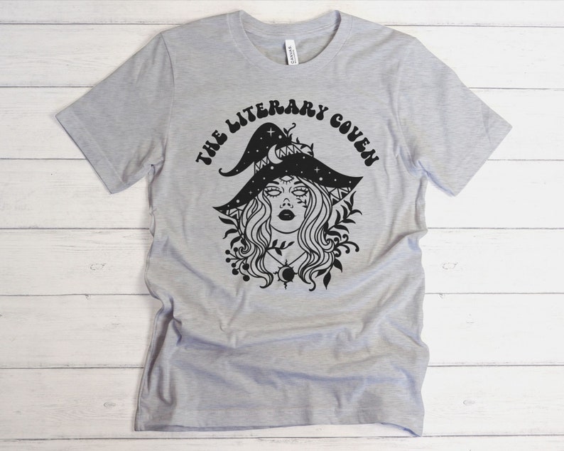 Peut inclure: Un t-shirt gris avec un motif noir d'une sorci&egrave;re portant un chapeau orn&eacute; d'&eacute;toiles et d'un croissant de lune. Le texte "The Literary Coven" est &eacute;crit dans une police r&eacute;tro autour de la sorci&egrave;re.
