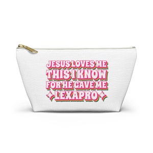 Op de afbeelding: Witte toilettas met rits met de tekst "Jesus loves me this I know for he gave me Lexapro" in roze en groene letters.