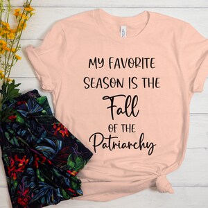 Op de afbeelding: Een perzikkleurig T-shirt met de tekst "My favorite season is the Fall of the Patriarchy" in zwart gedrukt.
