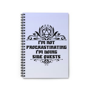 May include: A spiral bound notebook with a light purple cover featuring a black and white design of a 20-sided die with a decorative flourish and the text "I'm not procrastinating I'm doing side quests".