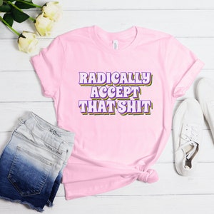 May include: Pink t-shirt with the text "RADICALLY ACCEPT THAT SHIT" in purple and yellow letters.
