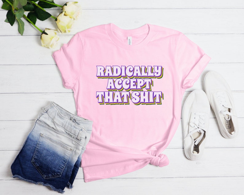 May include: Pink t-shirt with the text "RADICALLY ACCEPT THAT SHIT" in purple and yellow letters.