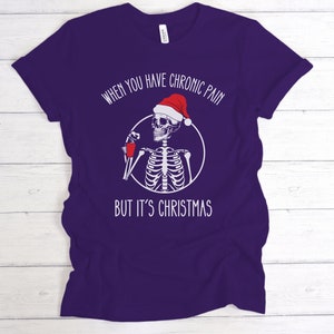 May include: Purple t-shirt with a white skeleton wearing a Santa hat and holding a red cup. The text on the shirt reads "When you have chronic pain but it's Christmas."