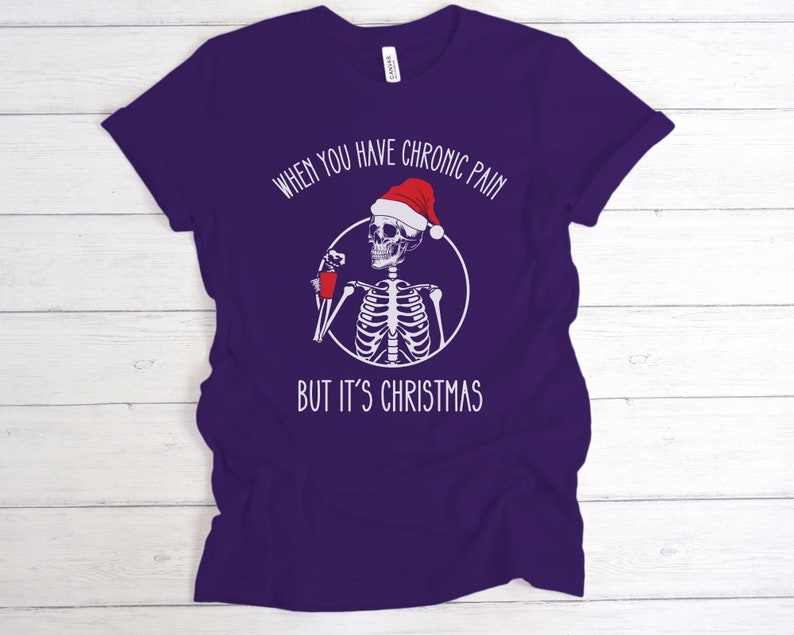 May include: Purple t-shirt with a white skeleton wearing a Santa hat and holding a red cup. The text on the shirt reads "When you have chronic pain but it's Christmas."