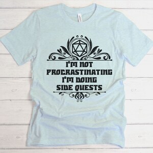 May include: Light blue t-shirt with the text "I'M NOT PROCRASTINATING I'M DOING SIDE QUESTS" in black, along with a decorative black design. The shirt is made of a soft material.