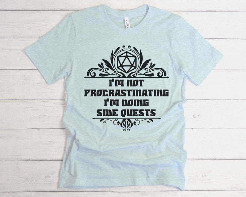May include: Light blue t-shirt with the text "I'M NOT PROCRASTINATING I'M DOING SIDE QUESTS" in black, along with a decorative black design. The shirt is made of a soft material.