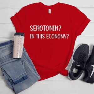 Pu&ograve; includere: Maglietta rossa con testo bianco che dice "SEROTONIN? IN THIS ECONOMY?"