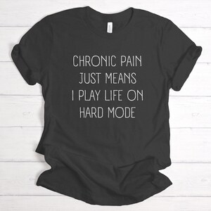 May include: Black t-shirt with white text that reads "Chronic pain just means I play life on hard mode."