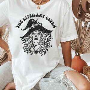 Peut inclure: T-shirt blanc avec un motif noir d'une sorci&egrave;re portant un chapeau orn&eacute; d'&eacute;toiles et d'un croissant de lune. Le texte "The Literary Coven" est &eacute;crit dans une police r&eacute;tro autour du motif.