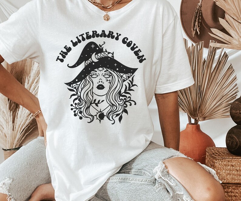 Peut inclure: T-shirt blanc avec un motif noir d'une sorci&egrave;re portant un chapeau orn&eacute; d'&eacute;toiles et d'un croissant de lune. Le texte "The Literary Coven" est &eacute;crit dans une police r&eacute;tro autour du motif.