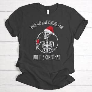 May include: Black t-shirt with a white skeleton wearing a Santa hat and holding a red cup. The text reads "When you have chronic pain but it's Christmas".