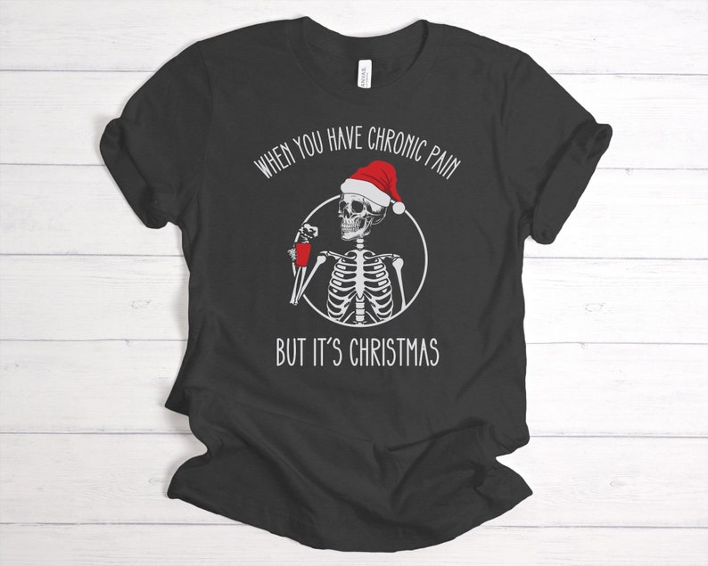 May include: Black t-shirt with a white skeleton wearing a Santa hat and holding a red cup. The text reads "When you have chronic pain but it's Christmas".