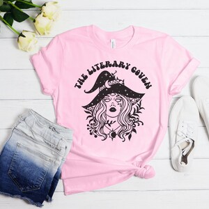 Peut inclure: T-shirt rose avec un graphique noir d'une sorci&egrave;re portant un chapeau orn&eacute; d'&eacute;toiles et d'un croissant de lune. Le texte "The Literary Coven" est &eacute;crit dans une police r&eacute;tro autour de la sorci&egrave;re.