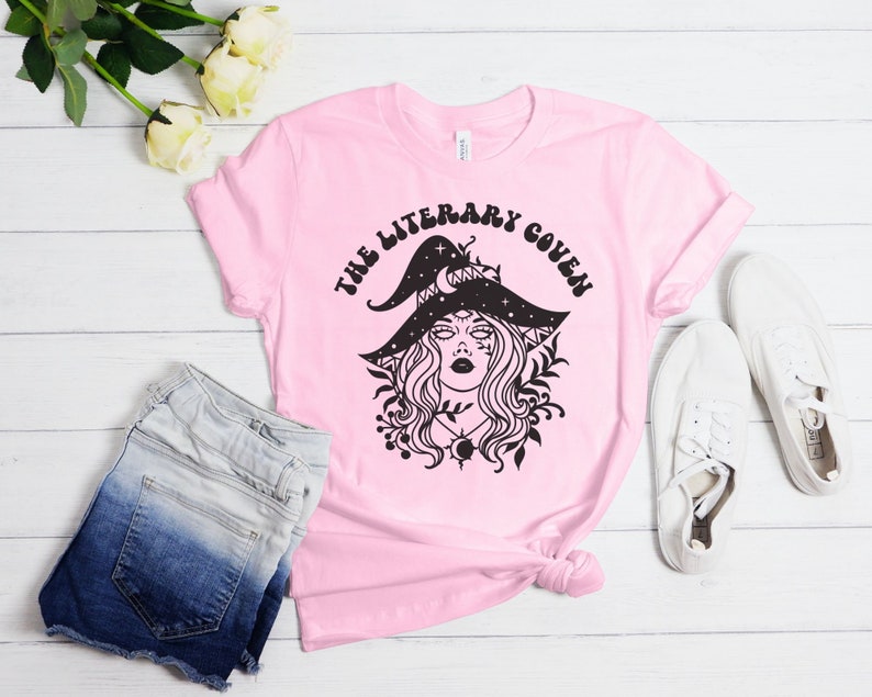 Peut inclure: T-shirt rose avec un graphique noir d'une sorci&egrave;re portant un chapeau orn&eacute; d'&eacute;toiles et d'un croissant de lune. Le texte "The Literary Coven" est &eacute;crit dans une police r&eacute;tro autour de la sorci&egrave;re.