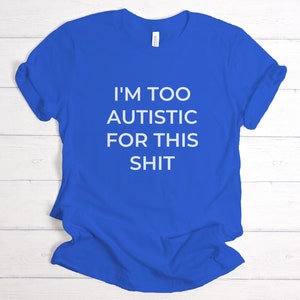 Puede incluir: Camiseta azul real con el texto blanco "I'M TOO AUTISTIC FOR THIS SHIT". La camiseta es de cuello redondo y parece estar hecha de un material suave. La camiseta est&aacute; doblada sobre una superficie de madera clara.