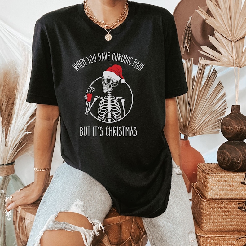 May include: Black t-shirt with a skeleton wearing a Santa hat and holding a red cup. The text on the shirt reads "When you have chronic pain but it's Christmas."