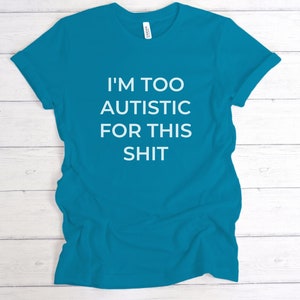 Puede incluir: Camiseta turquesa con el texto blanco "I'M TOO AUTISTIC FOR THIS SHIT". La camiseta de manga corta est&aacute; hecha de un material suave. La camiseta se muestra sobre una superficie de madera blanca.