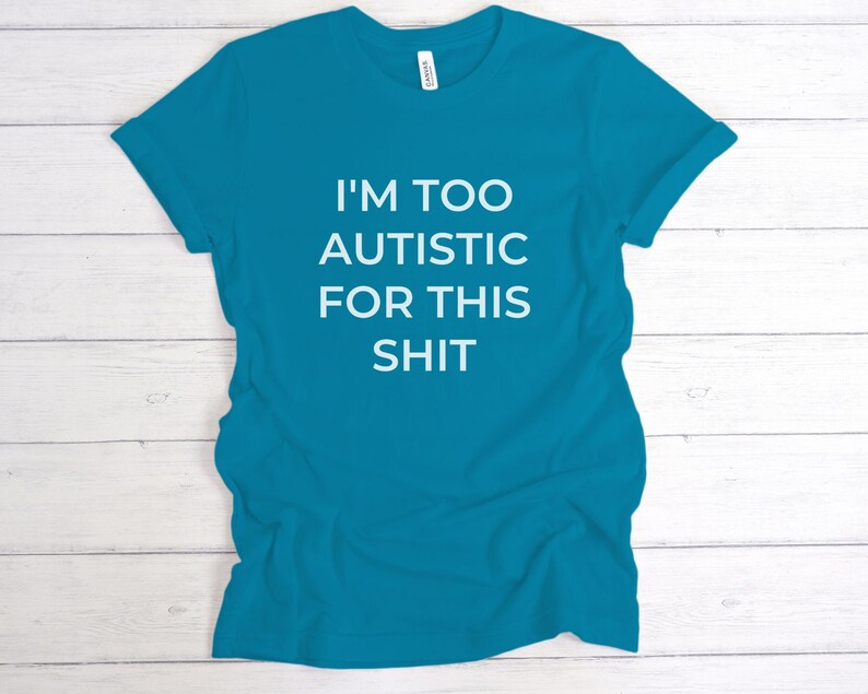Puede incluir: Camiseta turquesa con el texto blanco "I'M TOO AUTISTIC FOR THIS SHIT". La camiseta de manga corta est&aacute; hecha de un material suave. La camiseta se muestra sobre una superficie de madera blanca.