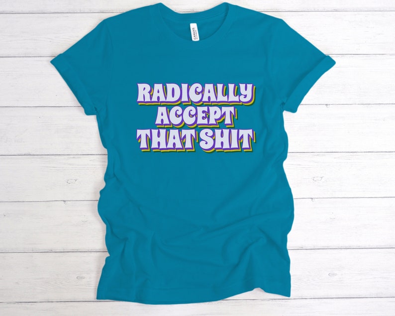 May include: Teal blue t-shirt with the text "RADICALLY ACCEPT THAT SHIT" in a retro font with purple and yellow outlines.