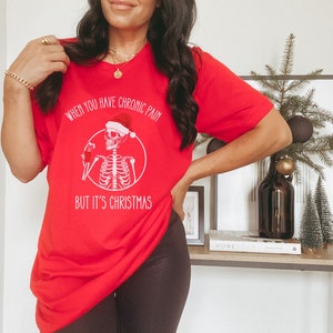 May include: Red Christmas themed t-shirt with a skeleton wearing a Santa hat and holding a drink. The text on the shirt reads "When you have chronic pain but it's Christmas."
