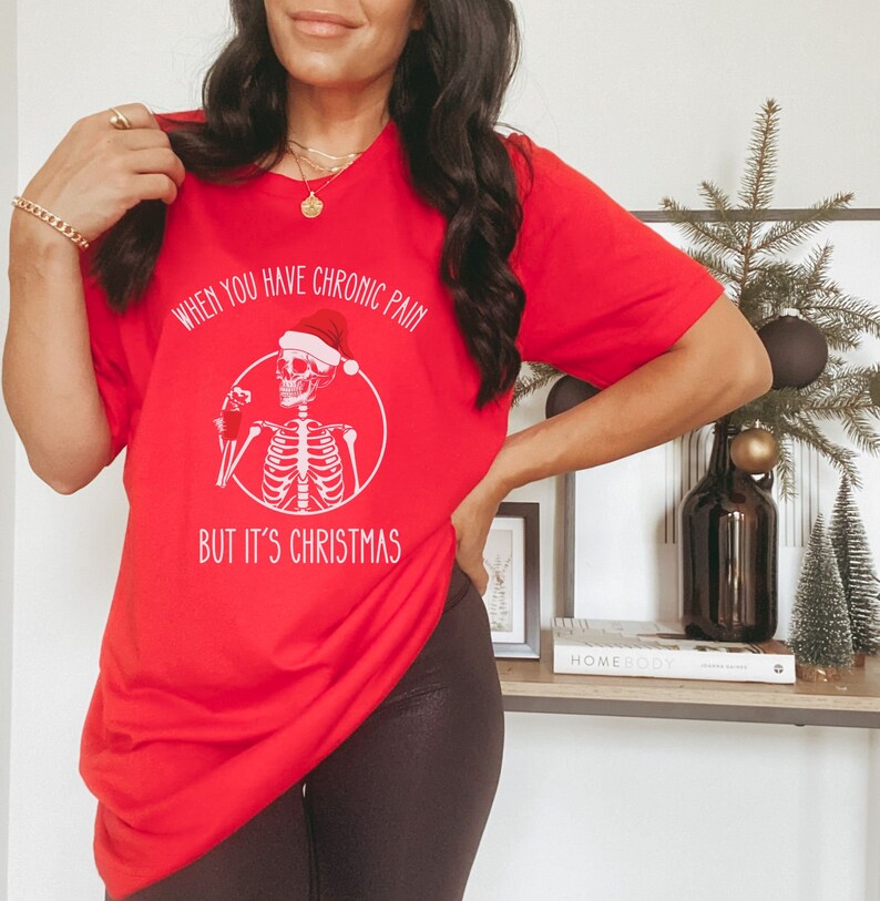 May include: Red Christmas themed t-shirt with a skeleton wearing a Santa hat and holding a drink. The text on the shirt reads "When you have chronic pain but it's Christmas."