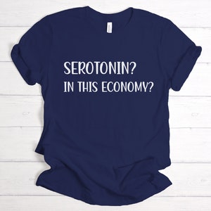 Pu&ograve; includere: Maglietta blu navy con testo bianco che dice "SEROTONIN? IN THIS ECONOMY?"
