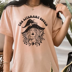Peut inclure: Un t-shirt rose p&acirc;le avec un motif noir d'une sorci&egrave;re portant un chapeau avec des &eacute;toiles et une lune croissante. Le texte "The Literary Coven" est &eacute;crit dans une police r&eacute;tro autour du motif.