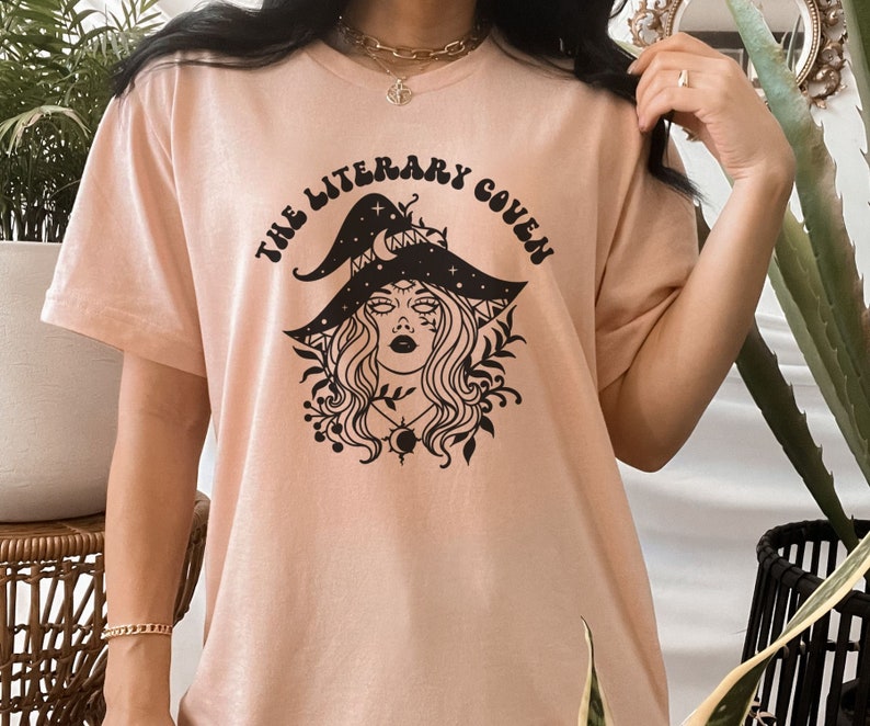 Peut inclure: Un t-shirt rose p&acirc;le avec un motif noir d'une sorci&egrave;re portant un chapeau avec des &eacute;toiles et une lune croissante. Le texte "The Literary Coven" est &eacute;crit dans une police r&eacute;tro autour du motif.