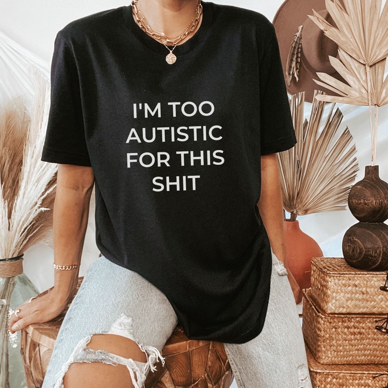 Puede incluir: Camiseta negra con el texto blanco "I'M TOO AUTISTIC FOR THIS SHIT". La camiseta es de cuello redondo. La persona lleva vaqueros claros rotos y joyas doradas.