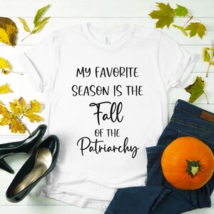 Op de afbeelding: Wit T-shirt met de tekst "My favorite season is the Fall of the Patriarchy" in zwart gedrukt.