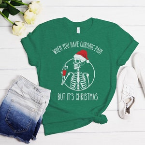 May include: Green t-shirt with a white skeleton wearing a Santa hat and holding a red cup. The text on the shirt reads "When you have chronic pain but it's Christmas."