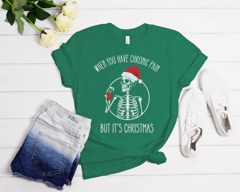 May include: Green t-shirt with a white skeleton wearing a Santa hat and holding a red cup. The text on the shirt reads "When you have chronic pain but it's Christmas."