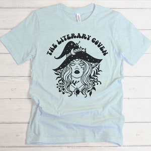 Peut inclure: T-shirt bleu clair avec un motif noir d'une sorci&egrave;re portant un chapeau avec des &eacute;toiles et une lune croissante. Le texte "The Literary Coven" est imprim&eacute; autour du motif.