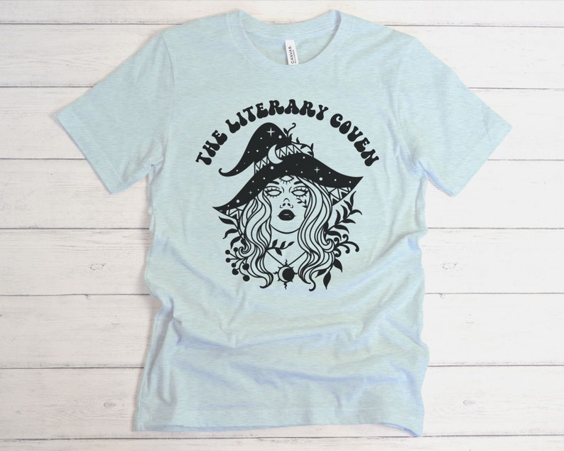 Peut inclure: T-shirt bleu clair avec un motif noir d'une sorci&egrave;re portant un chapeau avec des &eacute;toiles et une lune croissante. Le texte "The Literary Coven" est imprim&eacute; autour du motif.