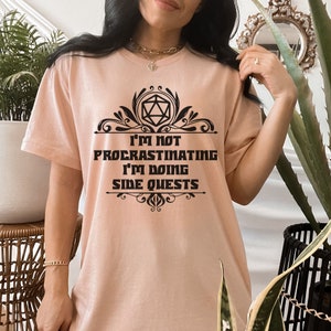 May include: A light peach-colored t-shirt with the text "I'M NOT PROCRASTINATING I'M DOING SIDE QUESTS" in black. The text is framed by decorative black flourishes and a geometric shape. The shirt is a casual wear item.