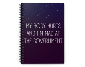 Può includere: Un quaderno a spirale di colore viola scuro e nero con un design di cielo notturno stellato. Il testo "MY BODY HURTS AND I'M MAD AT THE GOVERNMENT" è stampato in bianco sulla copertina.