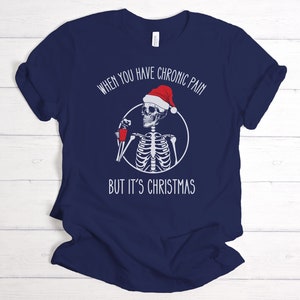 May include: Navy blue t-shirt with a white skeleton wearing a Santa hat and holding a red cup. The text on the shirt reads "When you have chronic pain but it's Christmas".